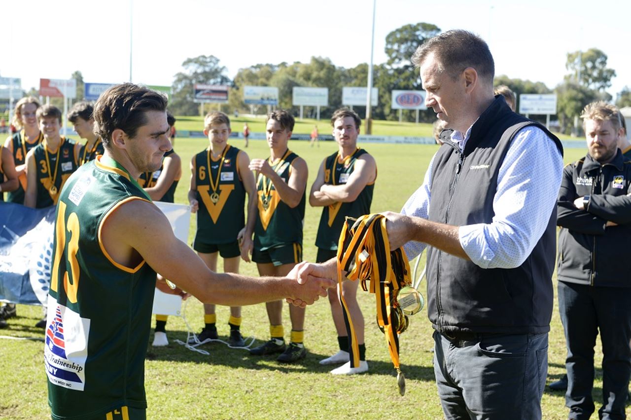 A man wearing a black vest jacket presents a medal to a football player in a green guernsey
