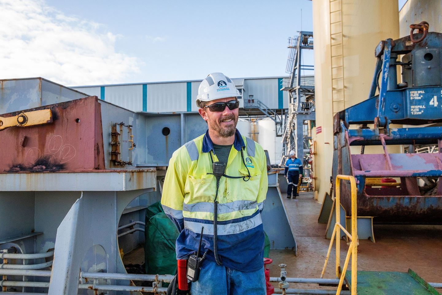 A man with a hard hat and a hi vis shirt stands on the deck of a ship, he is smiling