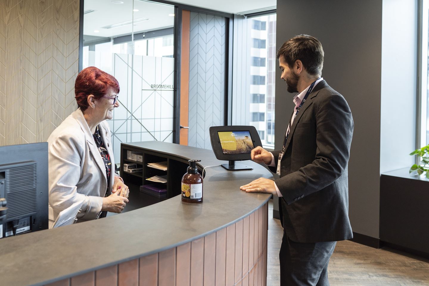 A woman with red hair stands behind a large curved desk, helping a man in a black suit to type on an iPad