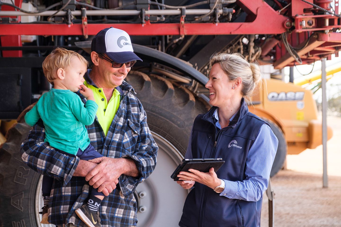 A blonde woman wearing a blue vest shows an ipad to a man wearing a checked shirt while he holds a toddler in his arms, they are both smiling.