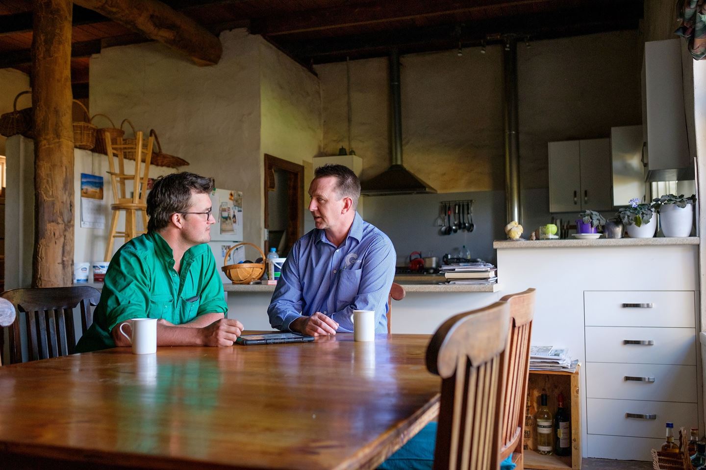 A man in a green shirt and a man in a blue shirt sit at a kitchen table chatting with coffee mugs in front of them