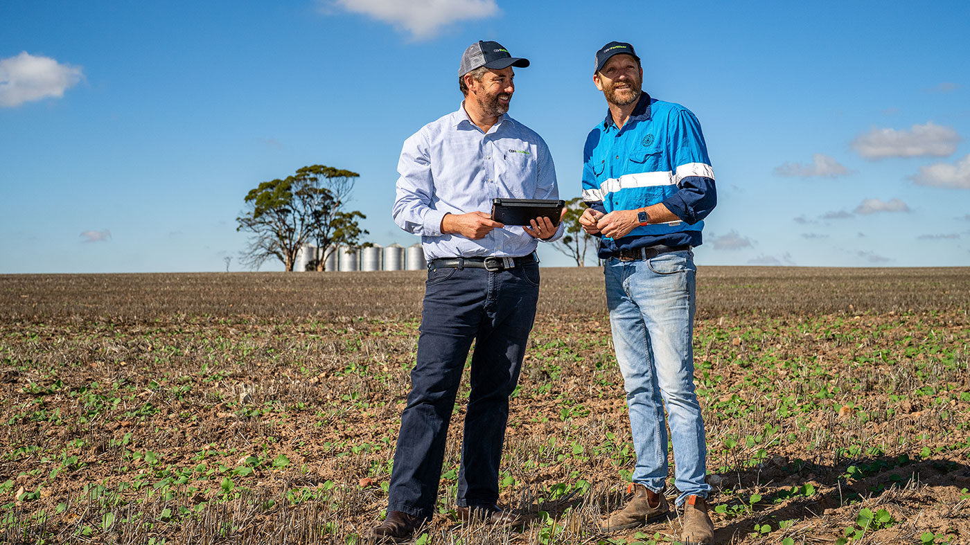 Two men wearing caps and workwear stand in a paddock looking at an ipad